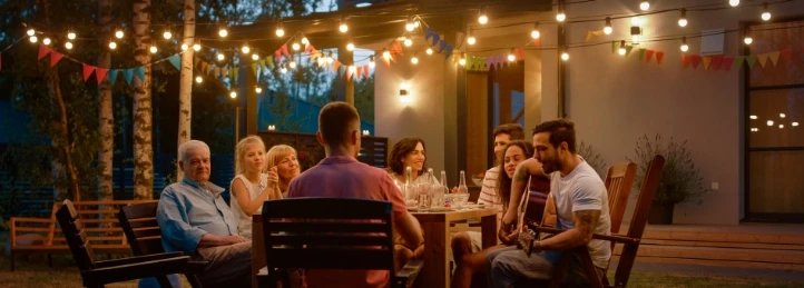 Family and friends gathered outdoors at night, sharing a meal under string lights on a backyard patio.
