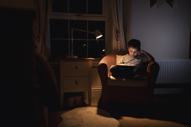 A young boy using a digital tablet while sitting on a chair. He is concentrating while playing on the tablet in a low lit room.